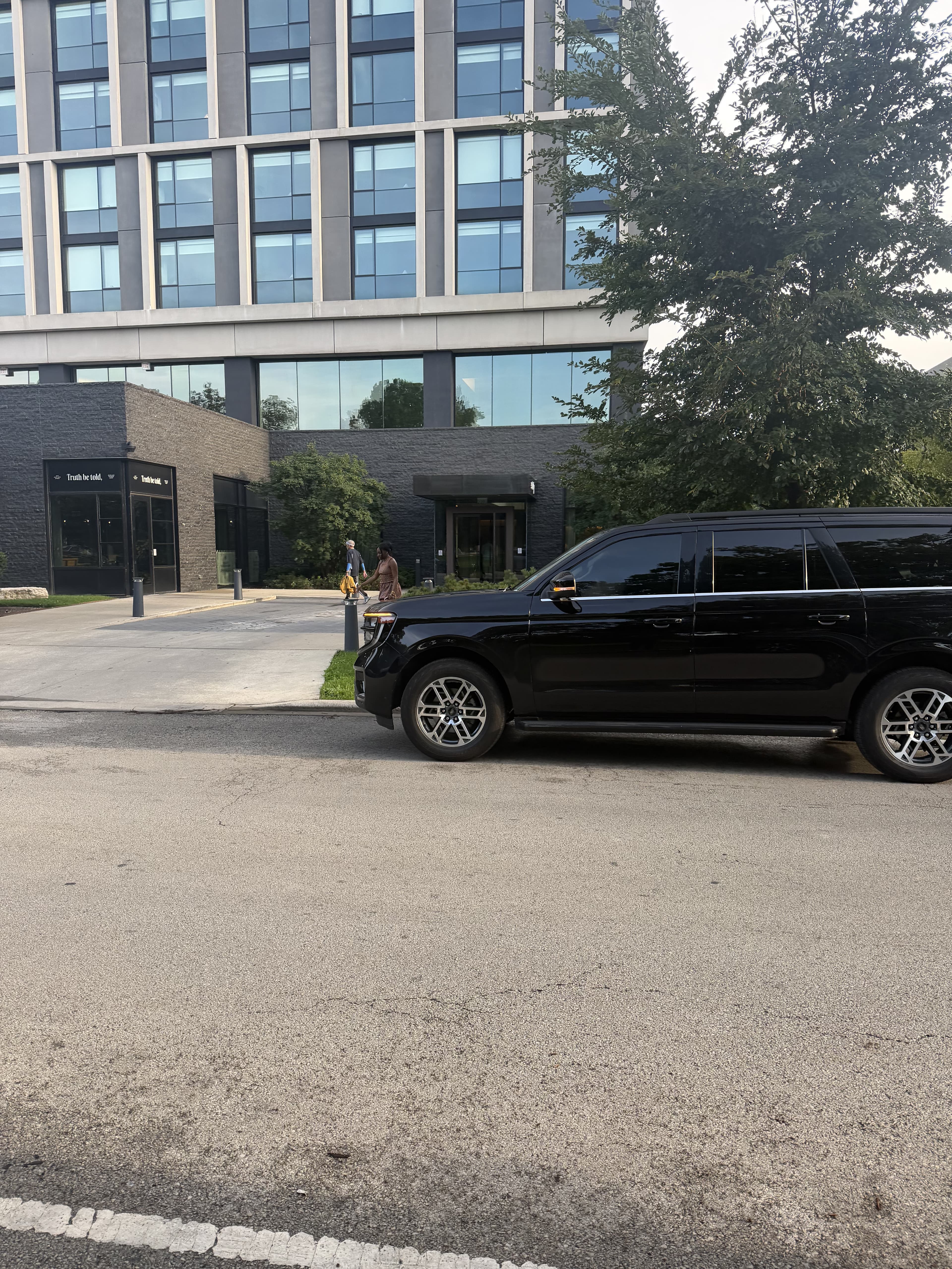 Black SUV parked on a street in front of a modern glass building with trees.
